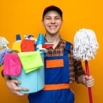 smiling-young-cleaning-guy-wearing-uniform-cap-holding-bucket-cleaning-tools-with-mop_141793-123193